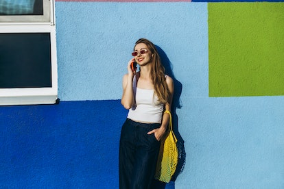 young woman talks on the phone as she leans against a blue wall and considers how the July 2023 supe...